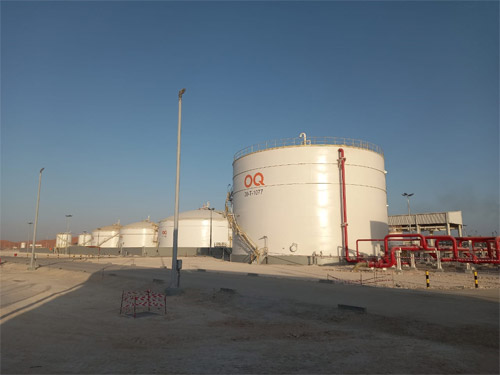 Large industrial storage tanks at OQ facility under a clear sky, with piping and safety equipment in the foreground.