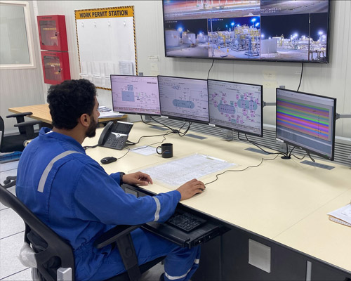 Engineer in blue coveralls monitoring multiple screens in a control room at an industrial plant or facility.