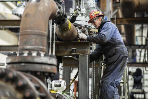 Engineer repairing industrial machinery, wearing safety gear and helmet, in a factory setting.