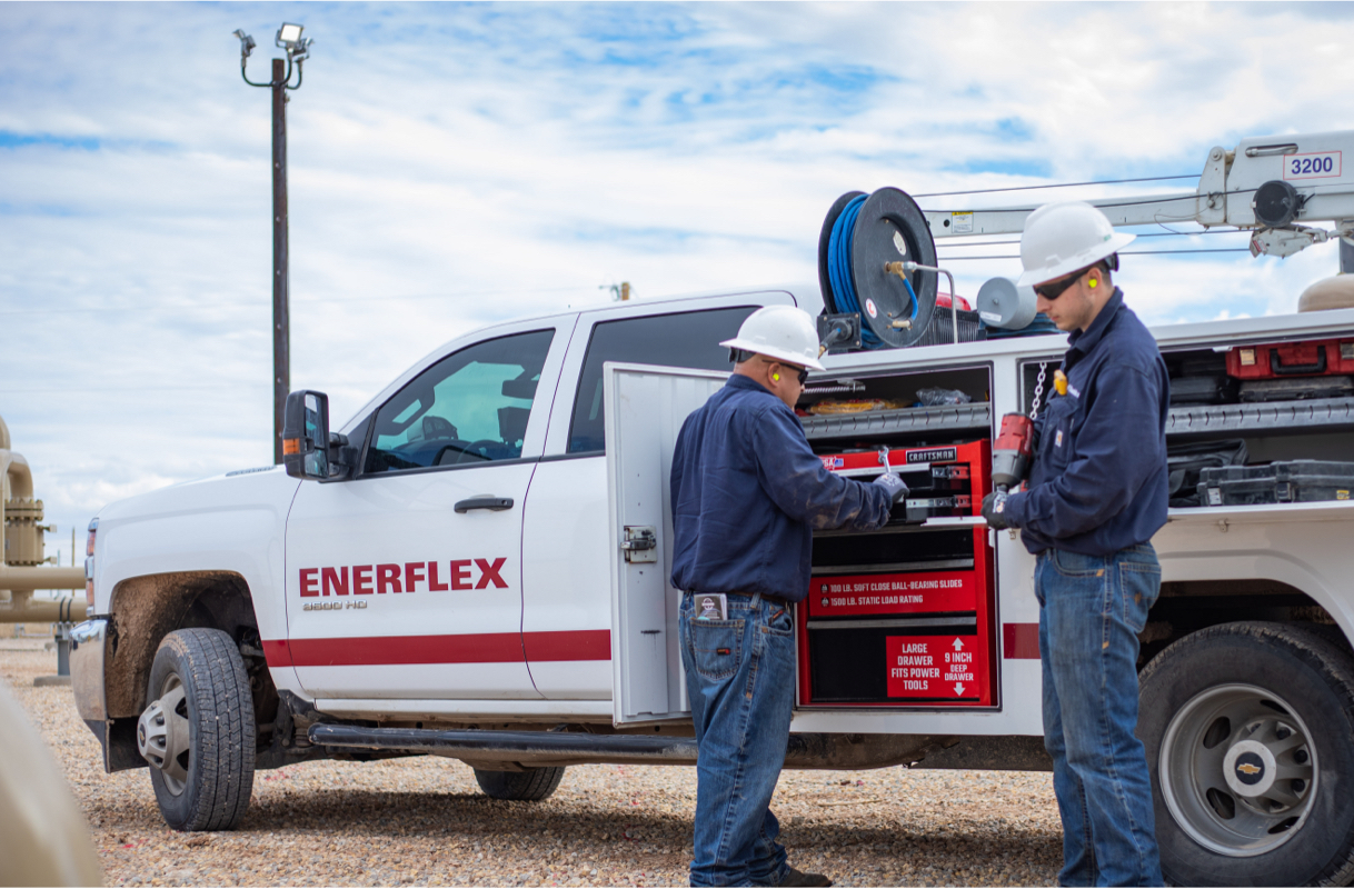 Two Enerflex field technicians work on a service truck equipped with tools and machinery in an industrial outdoor setting.